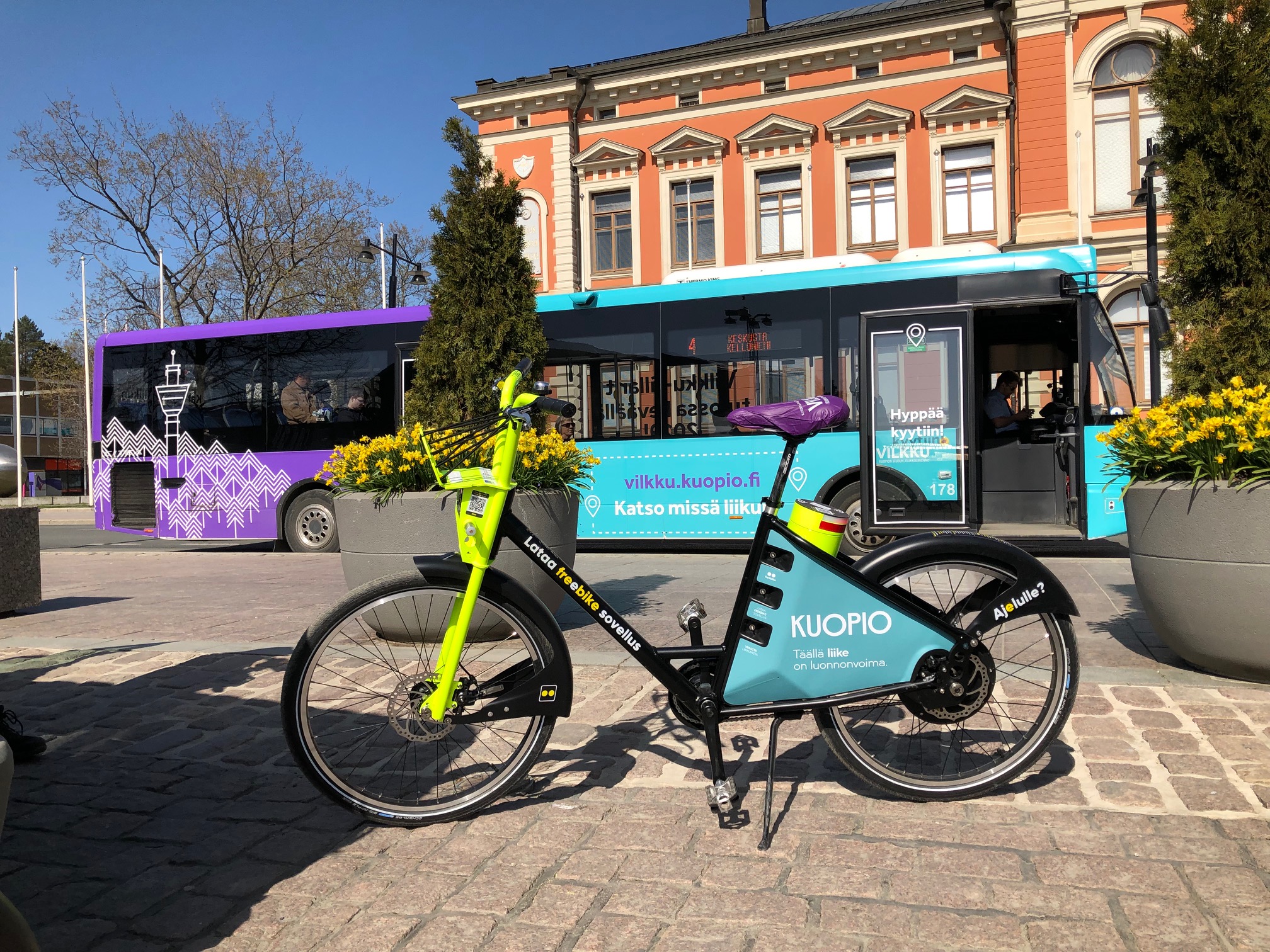 Kuvassa etualalla Vilkku-fillari ja taka-alalla Vilkun uusin väreihin teipattu bussi. Kuva on otettu aurinkoisena päivänä Kuopion kaupungintalon edessä.